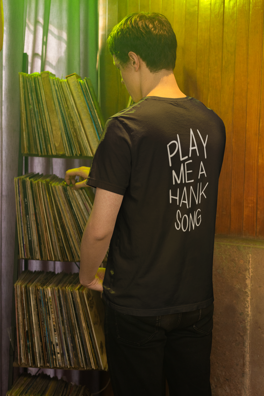 Black t-shirt with 'Play me a Hank song' text worn by a man browsing vinyl records in front of a record collection.